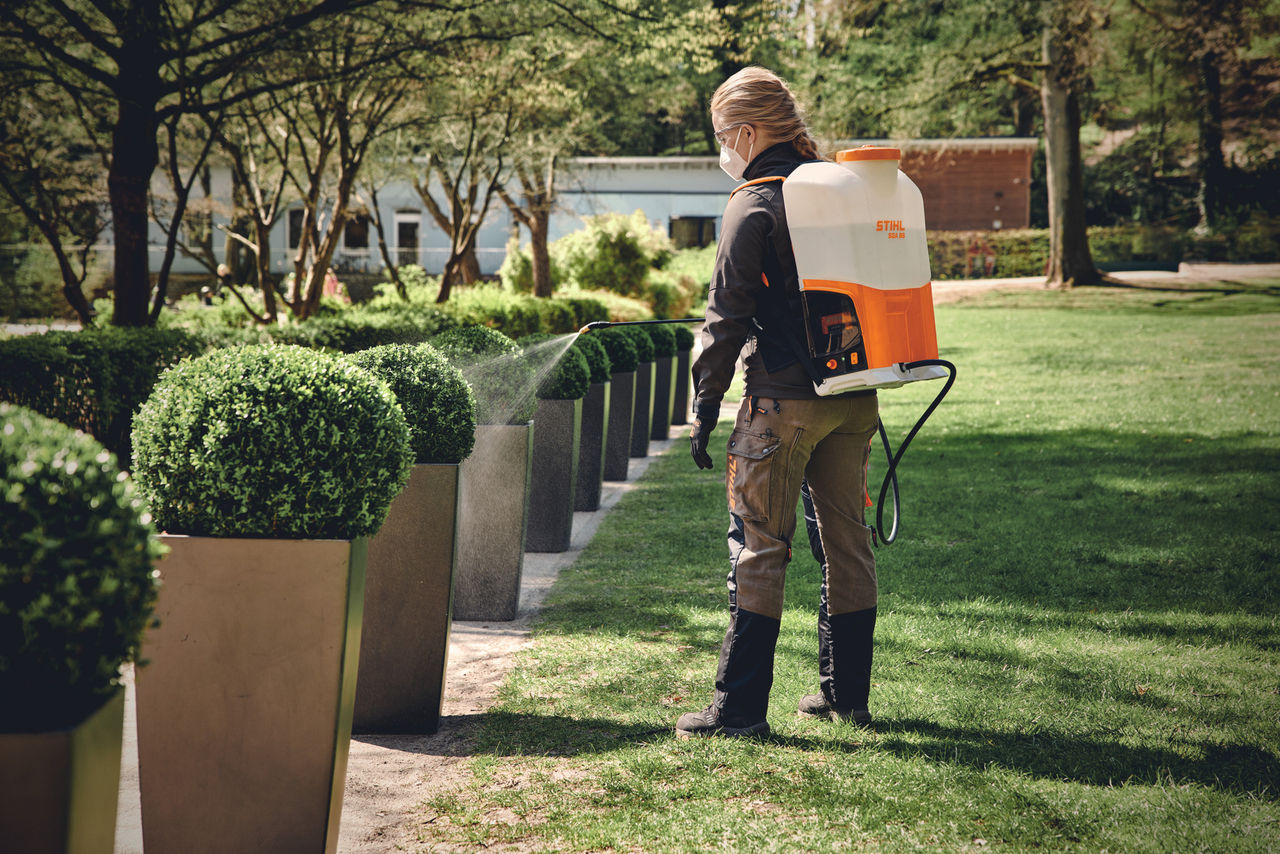 フェイスマスクをした金髪の男性が、STIHL 噴霧器を使って、緑地の小道に沿って丸く刈られた茂みに水をまく